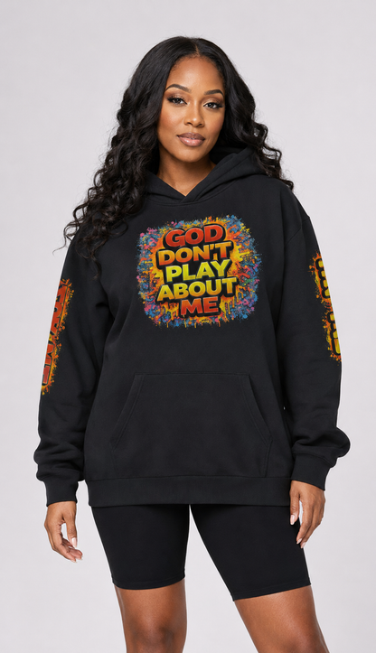A woman modeling a black pullover hoodie with a colorful graphic on the chest that reads 'GOD DON'T PLAY ABOUT ME' and matching graphics on the sleeves, paired with black biker shorts against a light gray studio background.